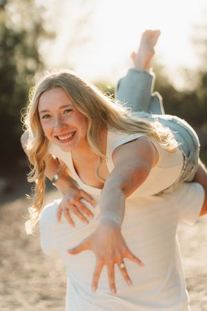 Smiling blonde woman being carried over someone's shoulder outdoors in golden sunlight.