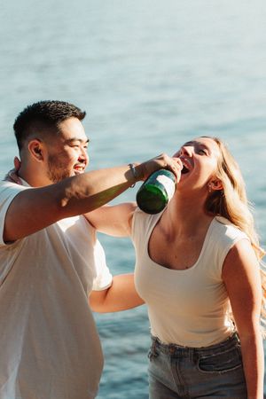A man playfully feeds a woman a sip from a green bottle near the water on a sunny day.