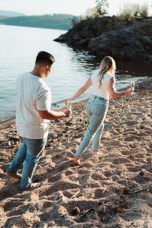 Couple walking barefoot on sandy beach holding champagne glasses near rocky shoreline at sunset.