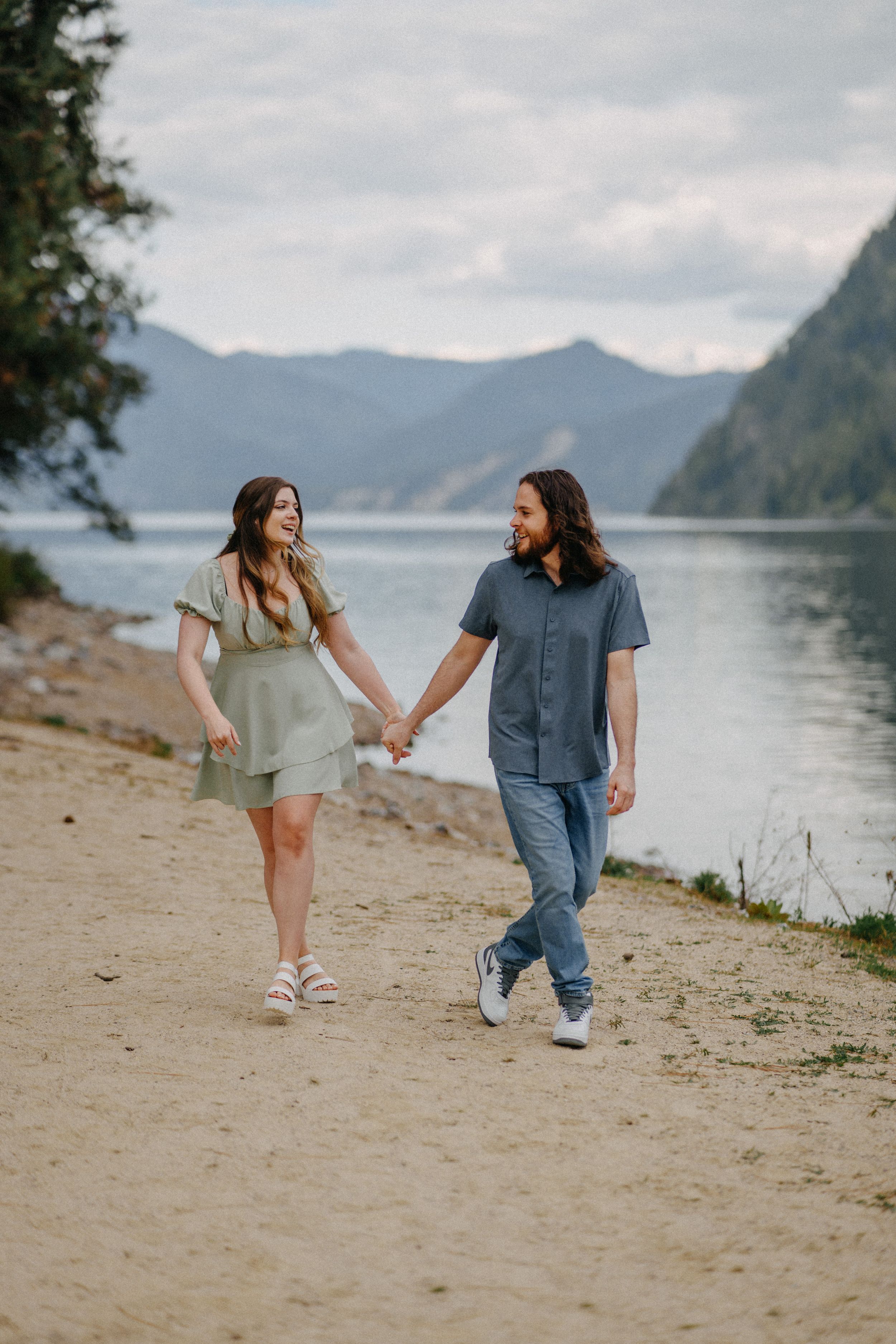 Couple holding hands walking along a sandy lakeside beach with misty mountains in the background.