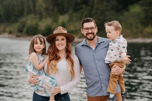 Happy family of four posing outdoors near a lake, parents holding young children, smiling in casual attire.