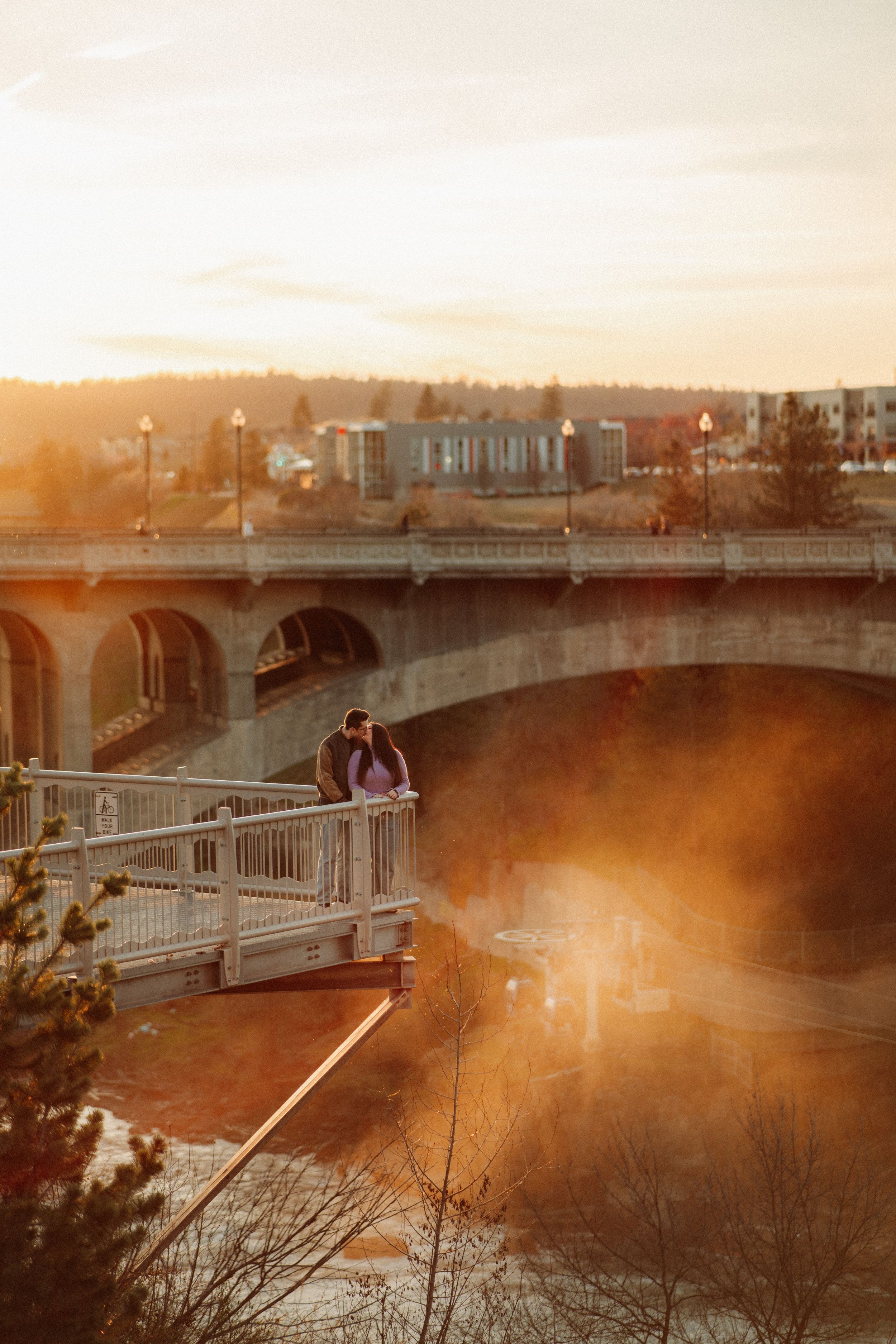 Couple embracing on a bridge at golden sunset with arched stone bridge and city skyline in background.
