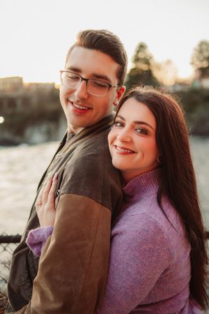 Young couple smiling at sunset, woman in pink sweater hugging man in glasses and brown jacket outdoors.