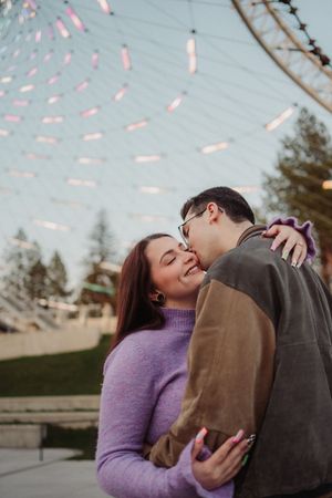 Couple sharing a romantic moment under colorful string lights at an outdoor venue, woman in purple sweater smiling.
