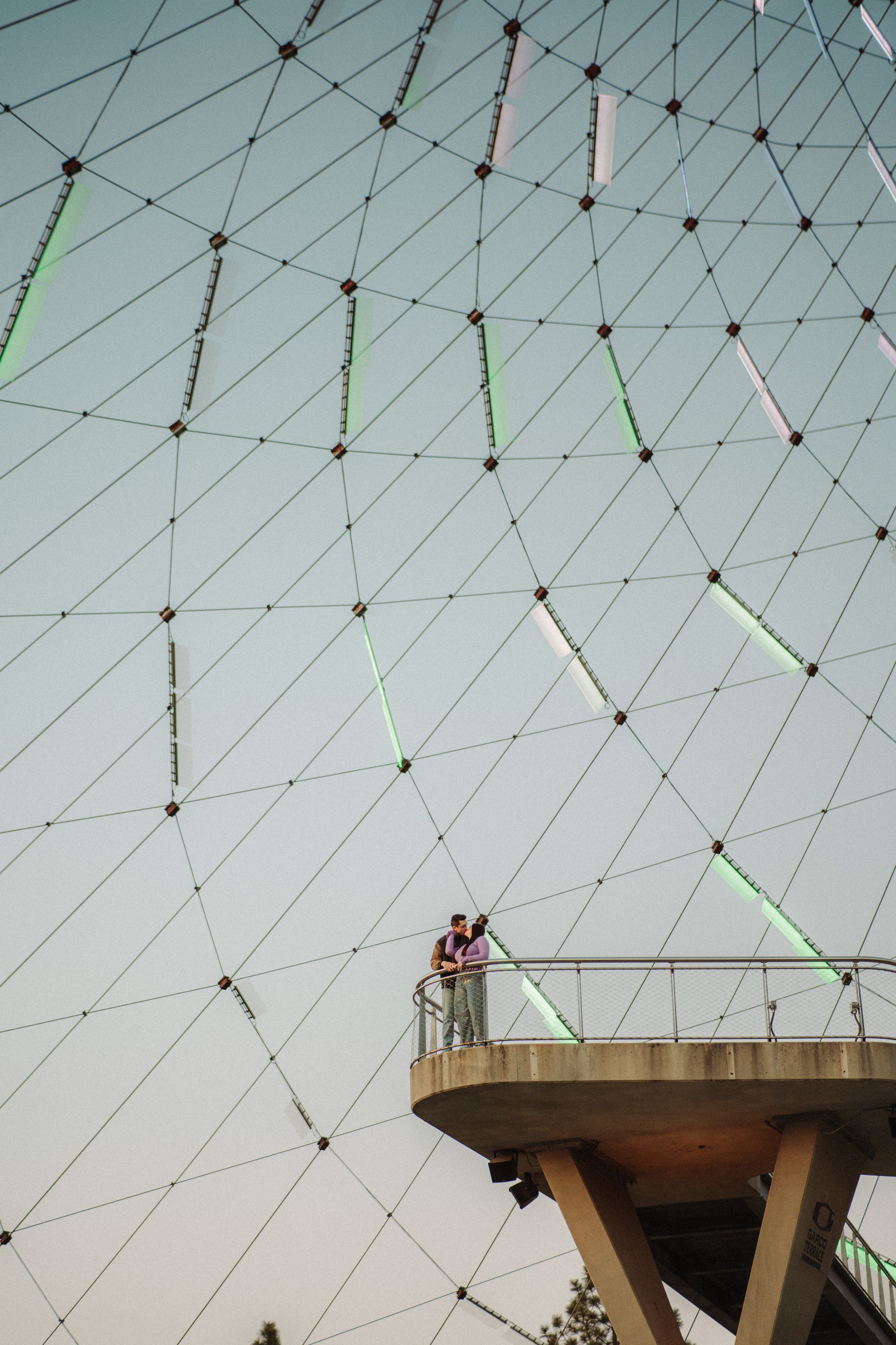 Two people stand on a elevated platform beneath a large geometric dome structure with a grey sky backdrop.