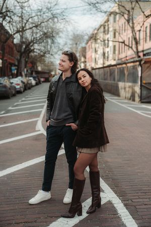 A stylish couple poses on an urban street, man in leather jacket and woman in brown coat with knee-high boots.