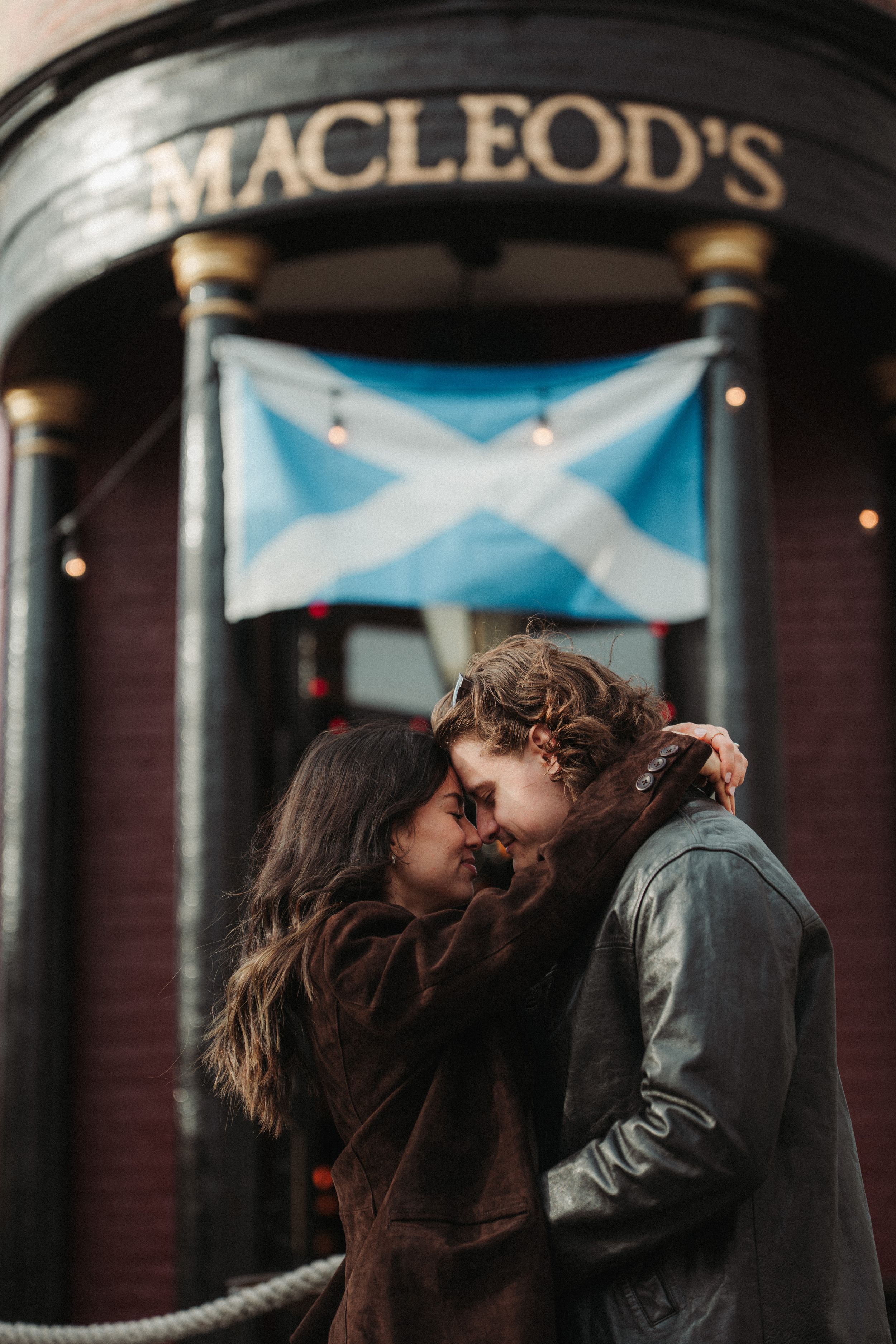 A romantic couple embraces outside Macleod's pub with a Scottish flag displayed above the entrance.