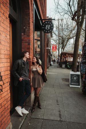 Couple smiling at each other on a brick-lined urban sidewalk, woman in brown skirt and boots, man in leather jacket.