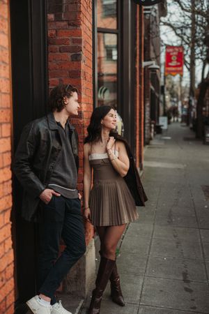 Young couple leaning against a brick building on a city sidewalk, woman in brown mini dress and boots, man in leather jacket.