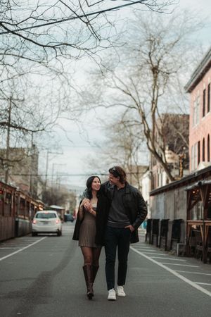 A couple walks arm in arm down a quiet urban street lined with bare trees and brick buildings on a cloudy day.