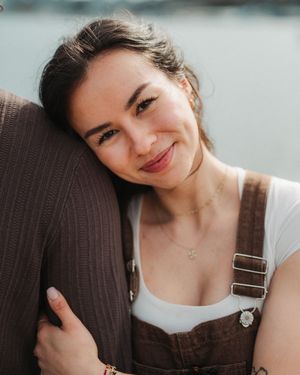 Young woman in brown overalls smiling warmly, leaning on someone's shoulder outdoors near water.