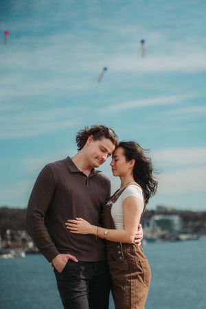 Romantic couple embracing near waterfront under blue sky with colorful kites, woman in overalls and man in brown sweater.