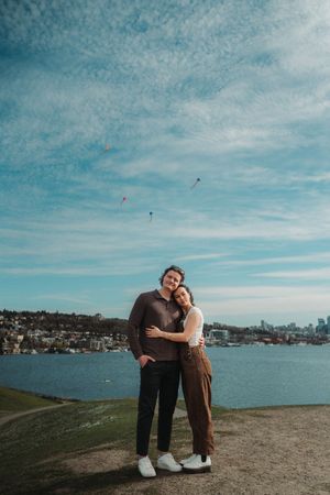 Couple embracing outdoors by a lake with kites flying in a blue sky, city skyline in the background.