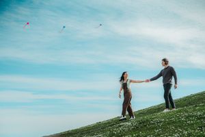 Couple holding hands on a grassy hill while colorful kites fly in a blue sky background.
