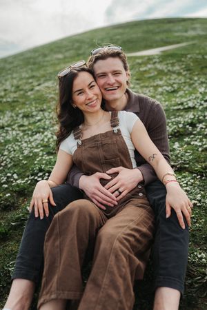 Happy couple sitting on a green hillside, woman in brown overalls leaning back into mans embrace, smiling outdoors.