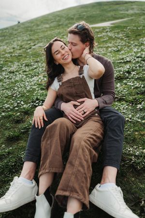 Couple sitting on a flower-covered hillside, man kissing woman on cheek while embracing her outdoors.