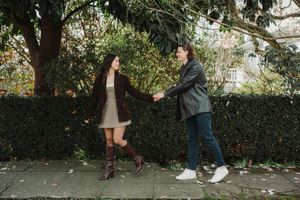 A couple holds hands and smiles while walking along a hedge-lined sidewalk surrounded by lush green trees.