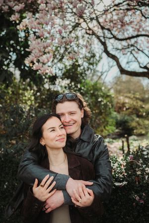 Couple embracing under blooming magnolia tree in spring garden, woman leaning back into mans arms, both smiling.
