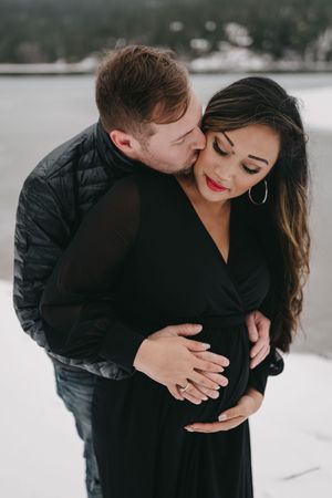Couple sharing a tender moment in snow, pregnant woman in black dress with red lips, man in black jacket embracing her belly.