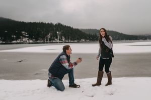 Man proposing to woman on snowy lakeside with frozen water and pine forest backdrop in winter.