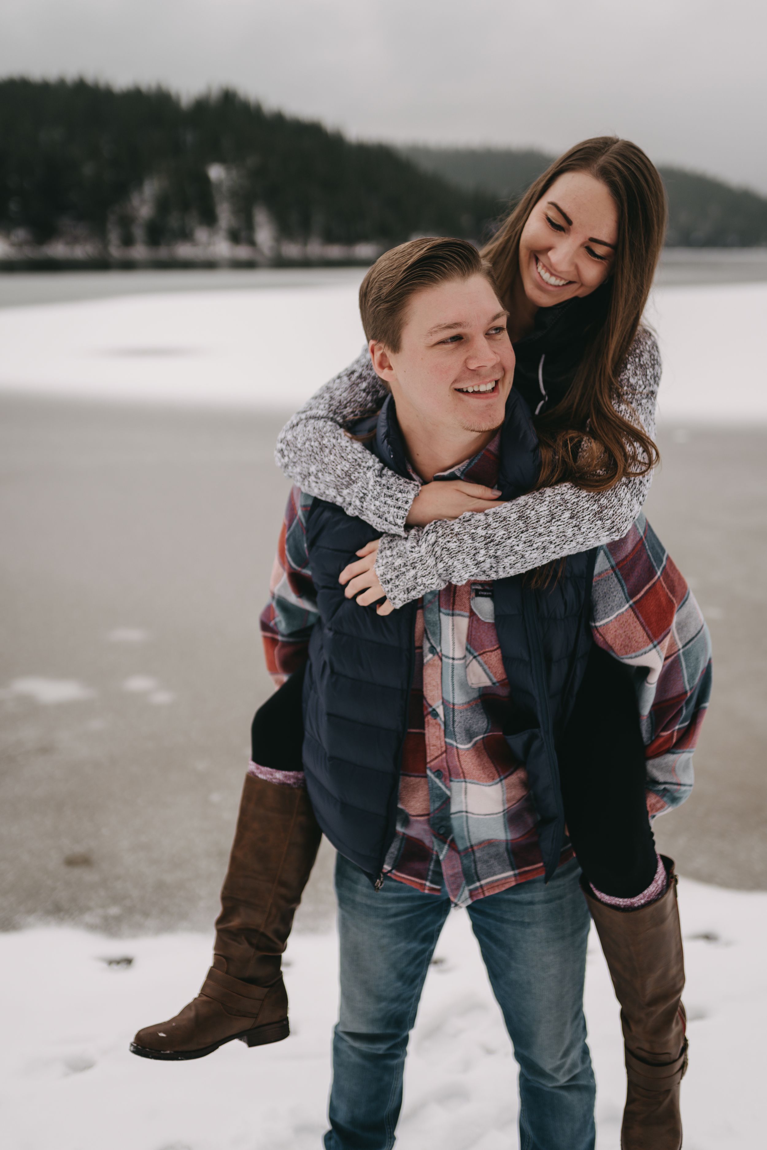 Smiling couple in winter, woman piggyback riding man near snowy frozen lake, wearing cozy plaid and knit outfits.