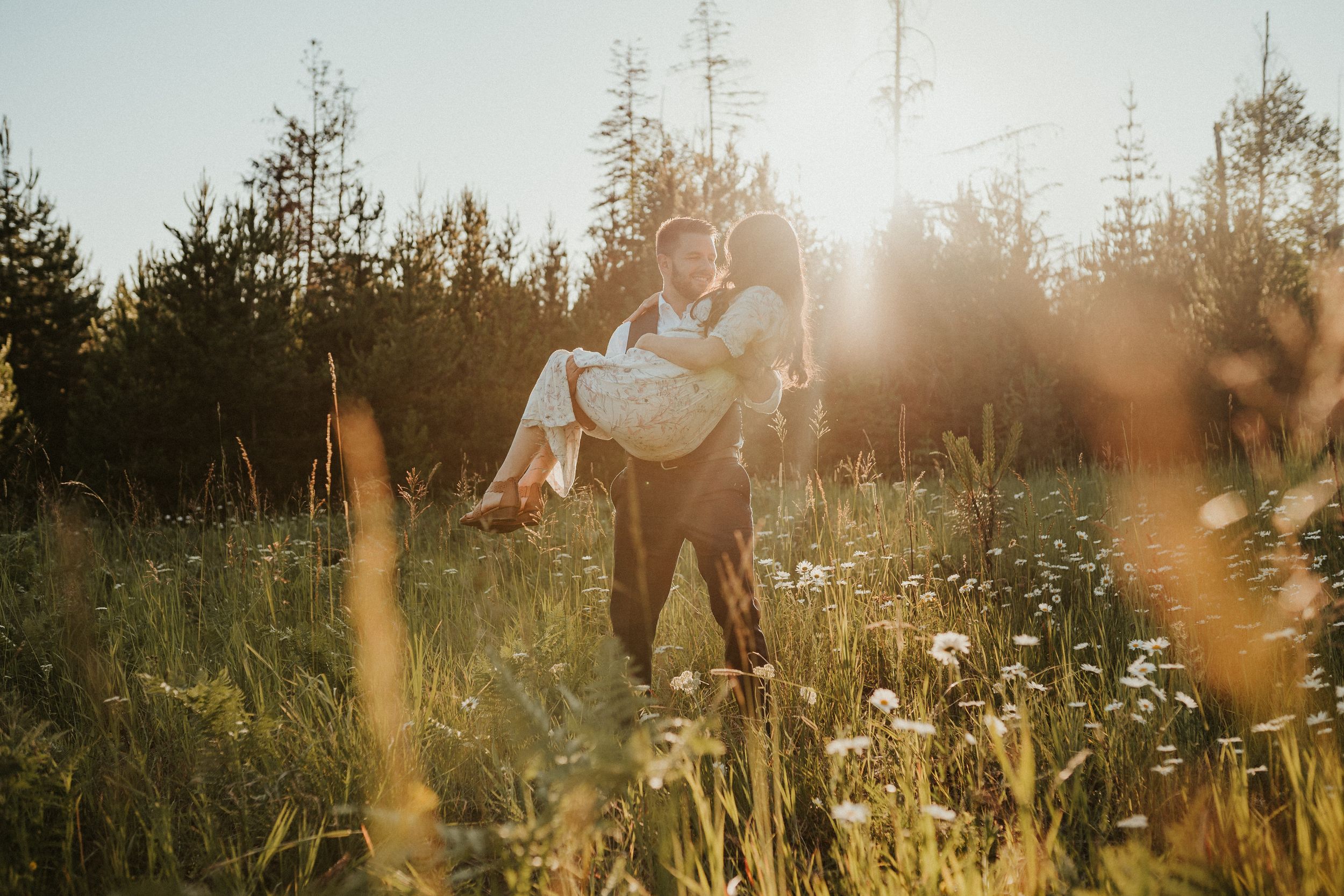 A couple embraces in a sunlit meadow surrounded by wildflowers and trees during golden hour.
