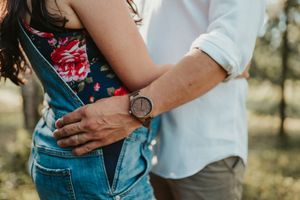 Couple embracing outdoors, man wearing a wooden watch, woman in floral top and denim overalls.