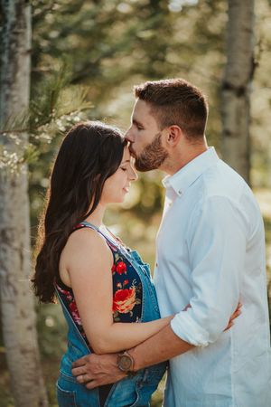A couple shares a tender forehead kiss outdoors among pine trees, woman in floral overalls and man in white shirt.