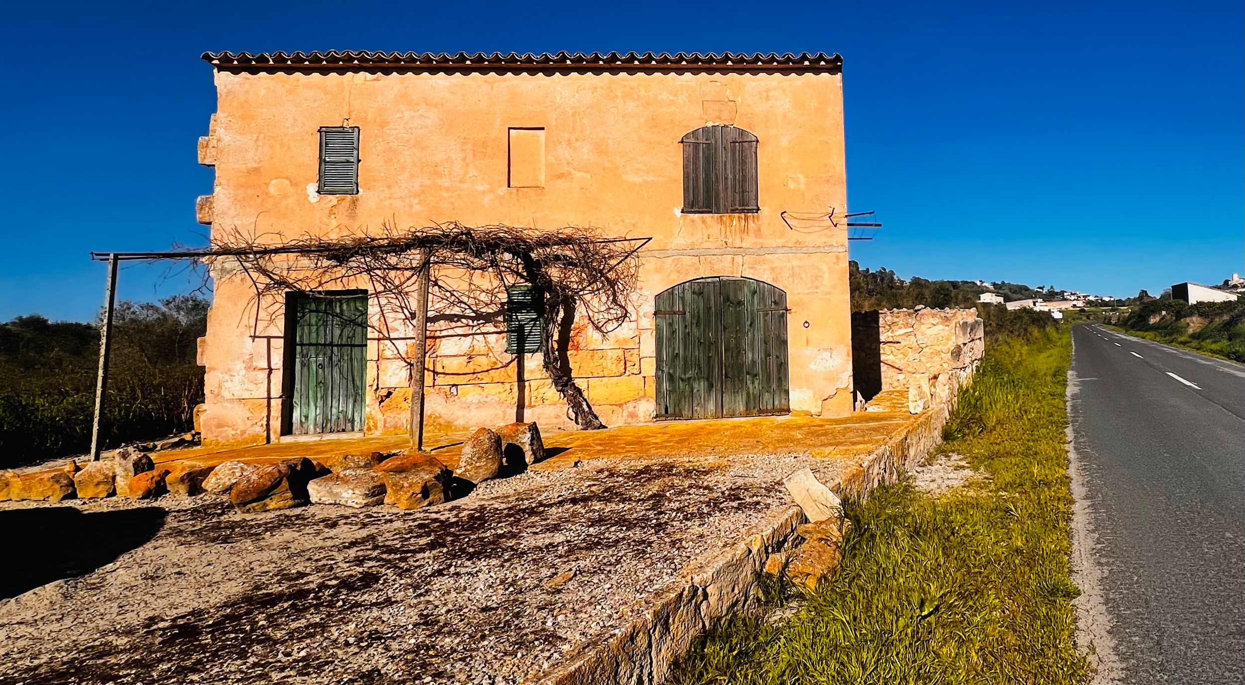 Rustic stone farmhouse with green shutters and climbing vines beside a rural road under a clear blue sky.