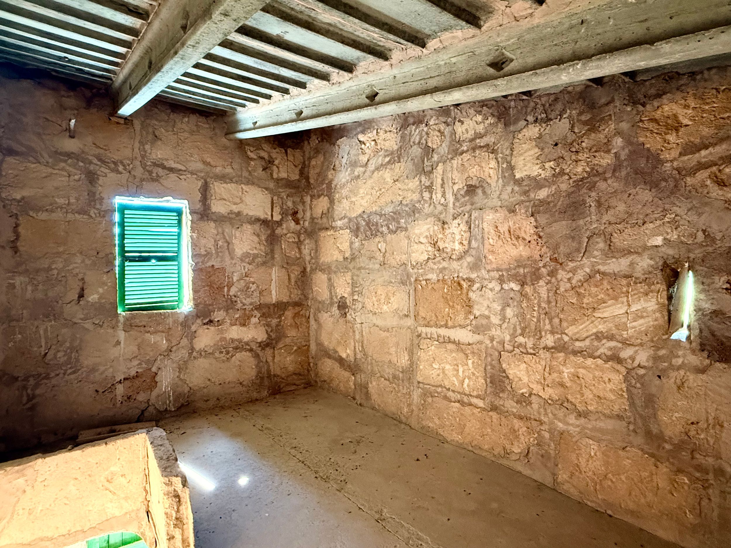 Interior stone wall room with green shuttered window, exposed wooden beam ceiling, and concrete floor.
