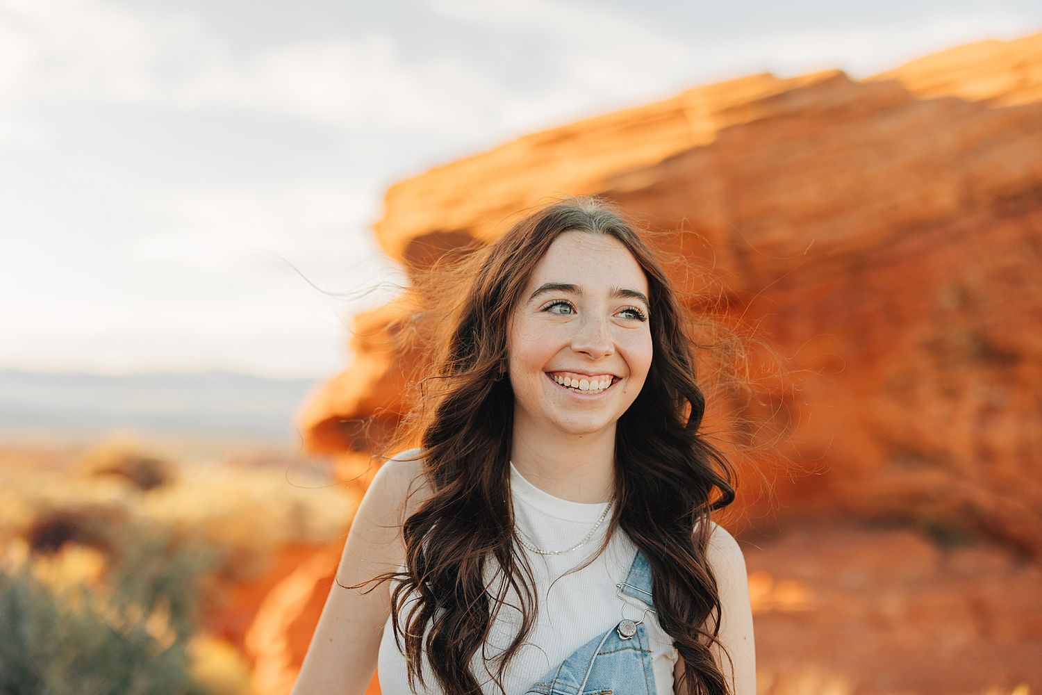 Smiling young woman with long wavy hair posing in front of red rock formations in a desert landscape.