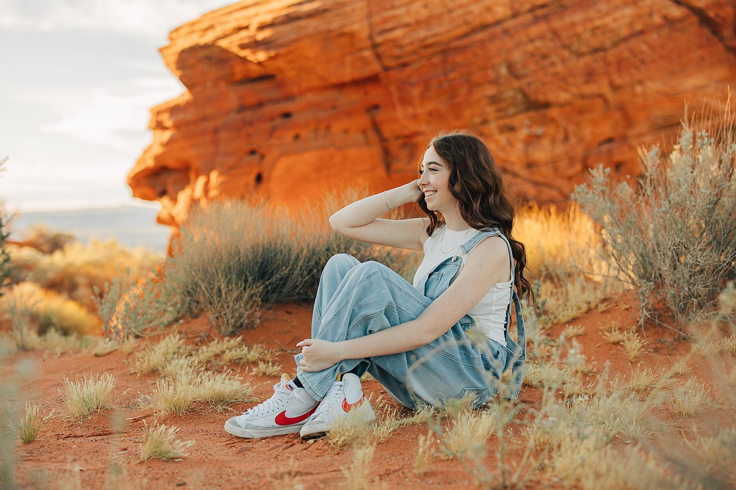 Young woman in overalls and Nike sneakers sitting on red desert sand near sandstone rocks at golden hour.