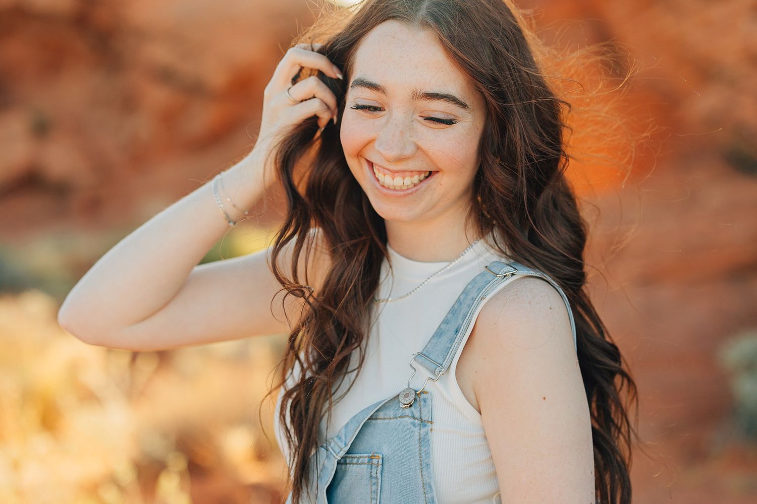 Smiling young woman with long wavy brown hair wearing denim overalls outdoors in a sunny desert setting.