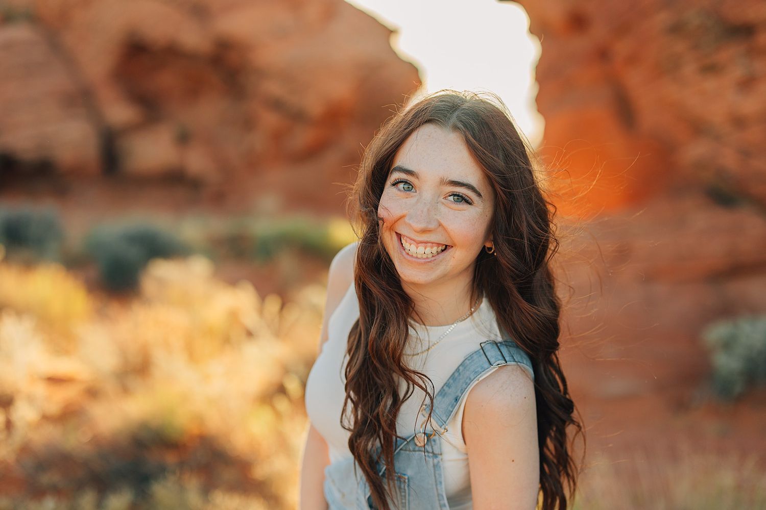 Young woman with long brown hair smiling outdoors at golden hour near red rock formations.