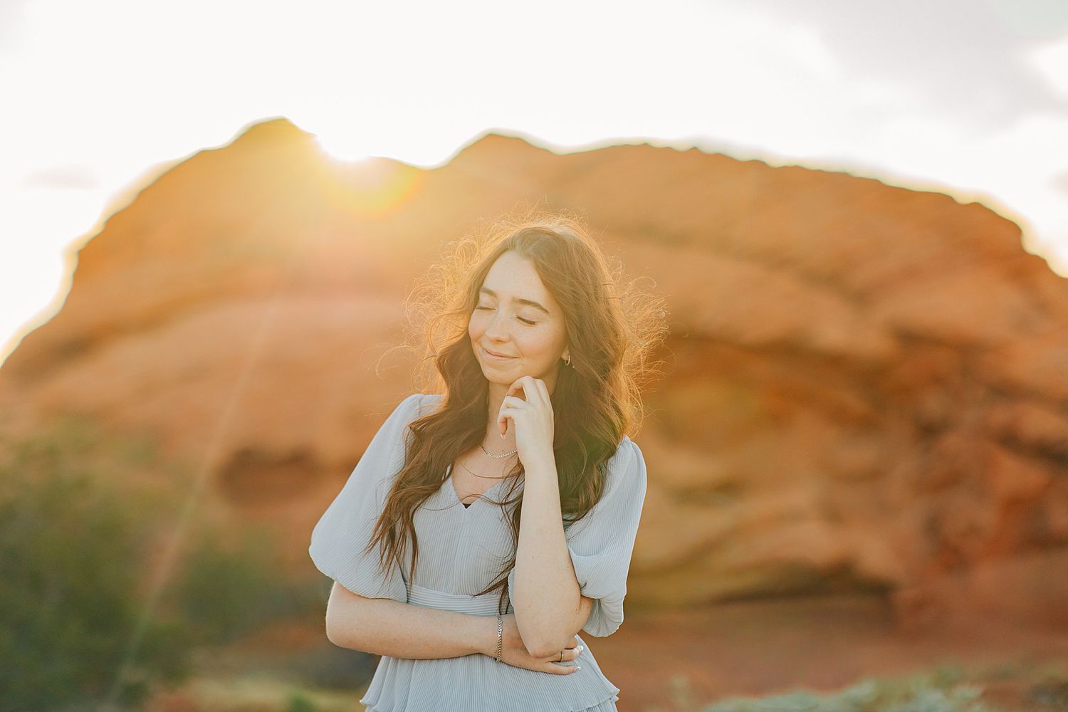 Young woman with long dark hair poses gracefully at golden hour near red rock formations in a serene outdoor setting.