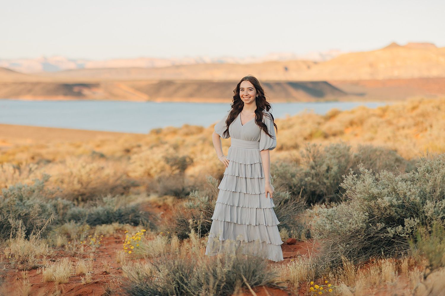 Woman in gray tiered maxi dress poses in desert landscape with lake and mountains at golden hour.