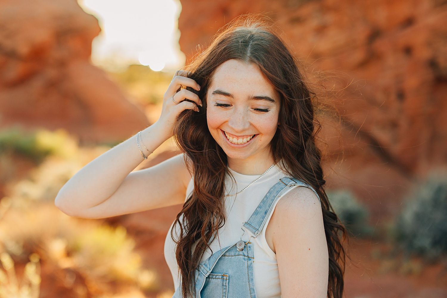 Smiling young woman with long wavy hair in denim overalls poses outdoors near red rock formations in golden sunlight.