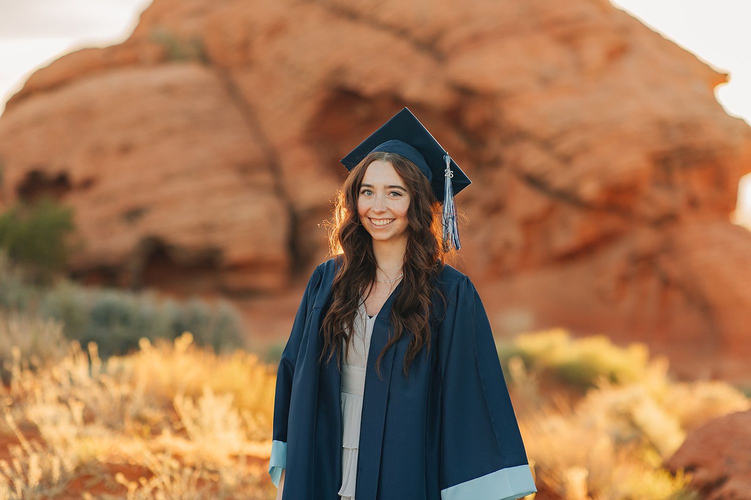 Young woman in navy graduation cap and gown smiling outdoors with red rock formations in background.