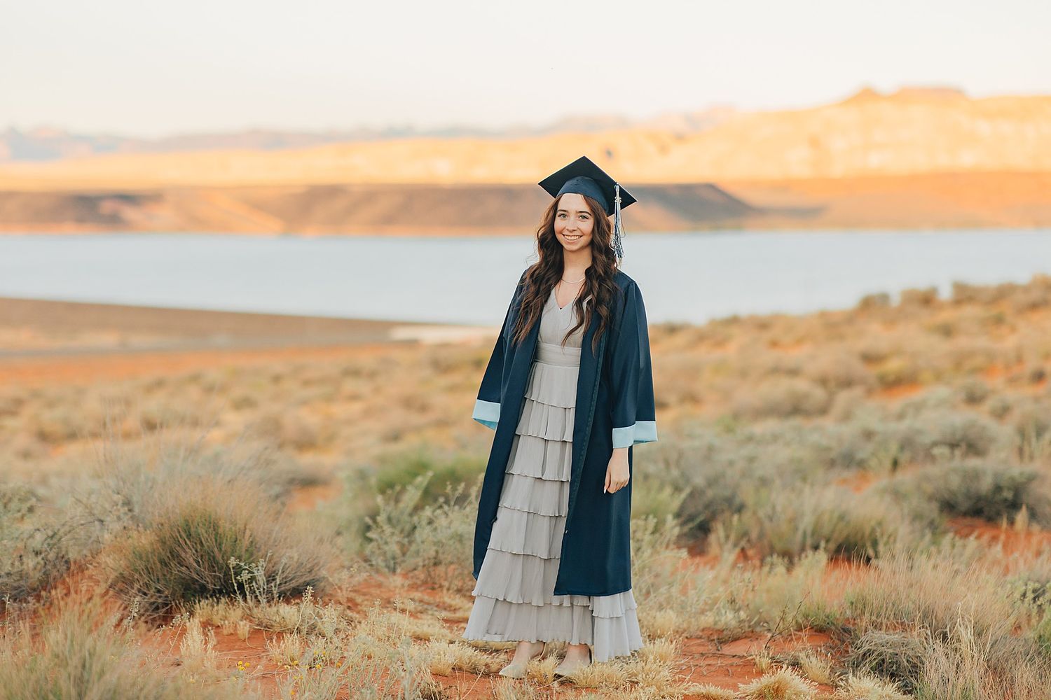 A smiling graduate in navy cap and gown poses in a desert landscape with a lake and mountains at sunset.