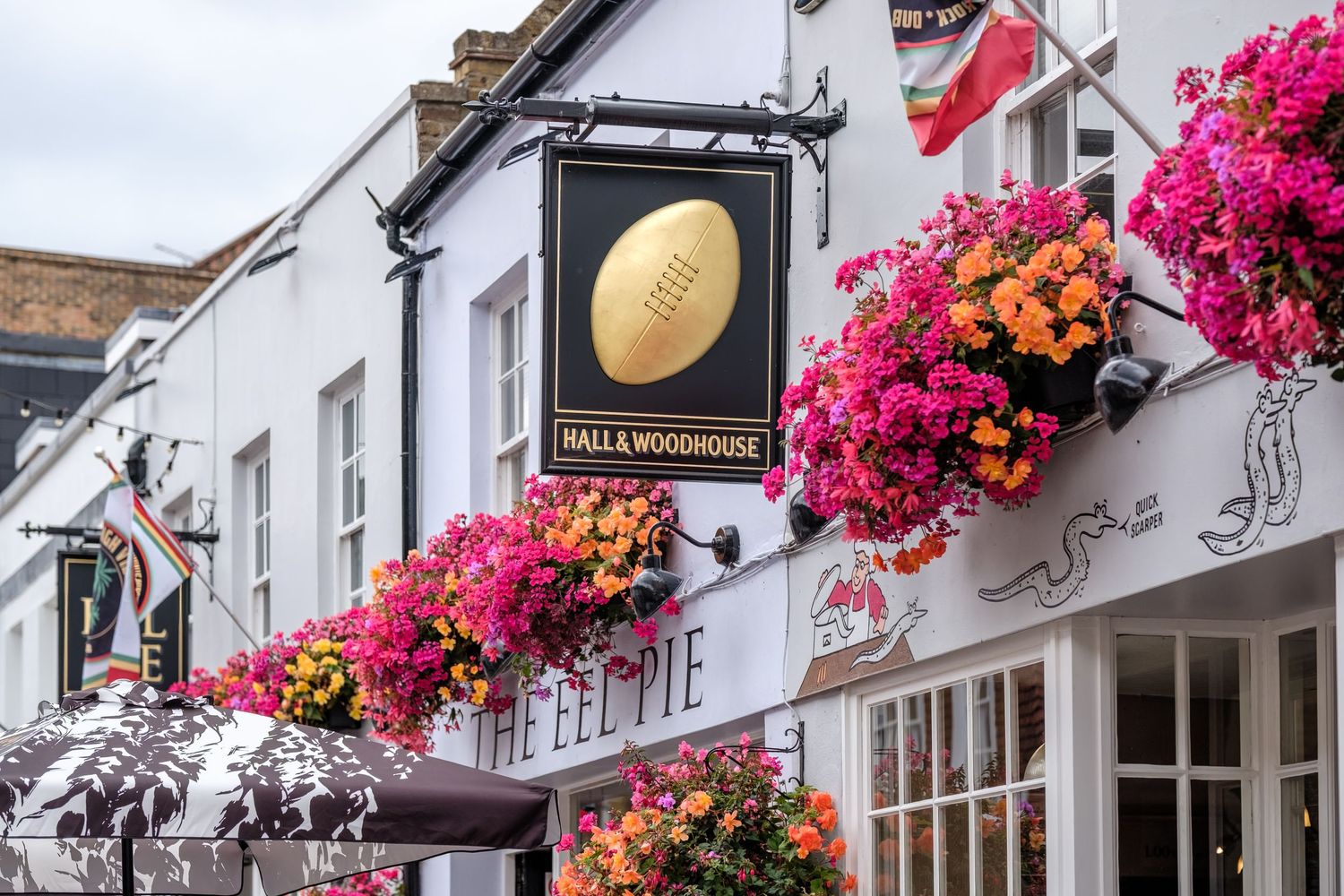 Hall & Woodhouse pub sign with golden rugby ball, adorned with vibrant pink and orange flower baskets on a charming street.