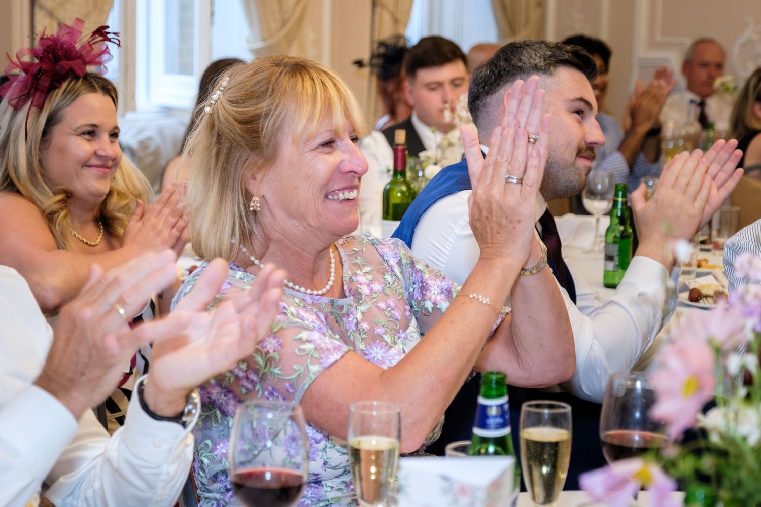 Guests clapping and smiling at a wedding reception dinner, dressed in formal attire at elegantly decorated tables.