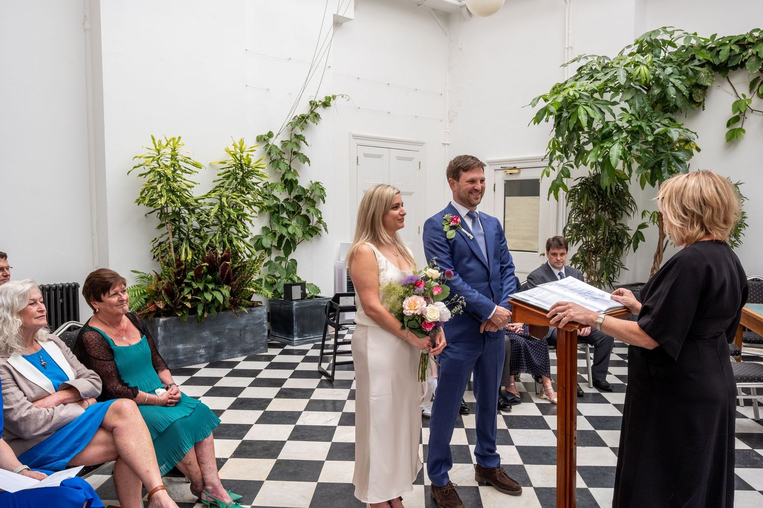 Couple exchanges wedding vows at indoor ceremony with checkered floor, officiant, and lush greenery backdrop.