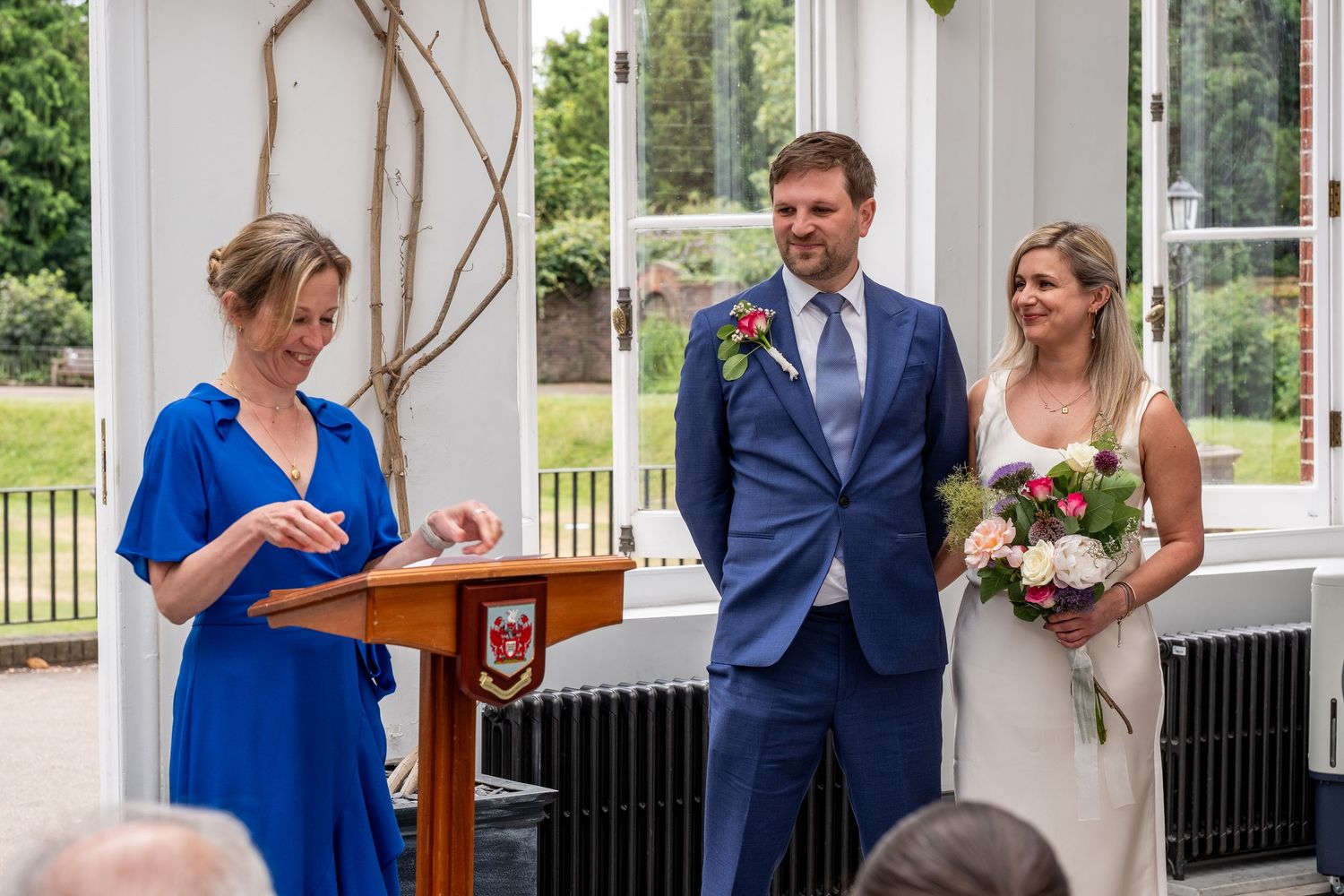 A wedding ceremony with a bride holding flowers and groom in blue suit standing before an officiant at a podium indoors.