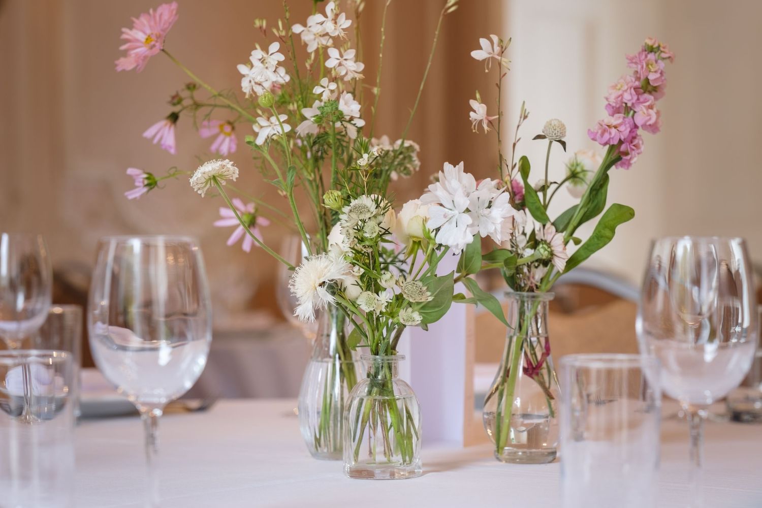 Elegant wildflower centerpieces in glass vases with pink and white blooms on a white tablecloth at a wedding reception.