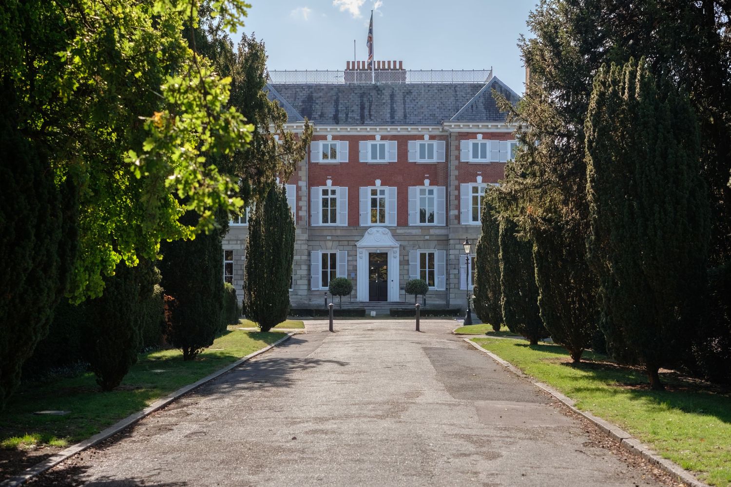 Grand red brick manor house with symmetrical facade, flanked by tall trees lining a wide gravel driveway.