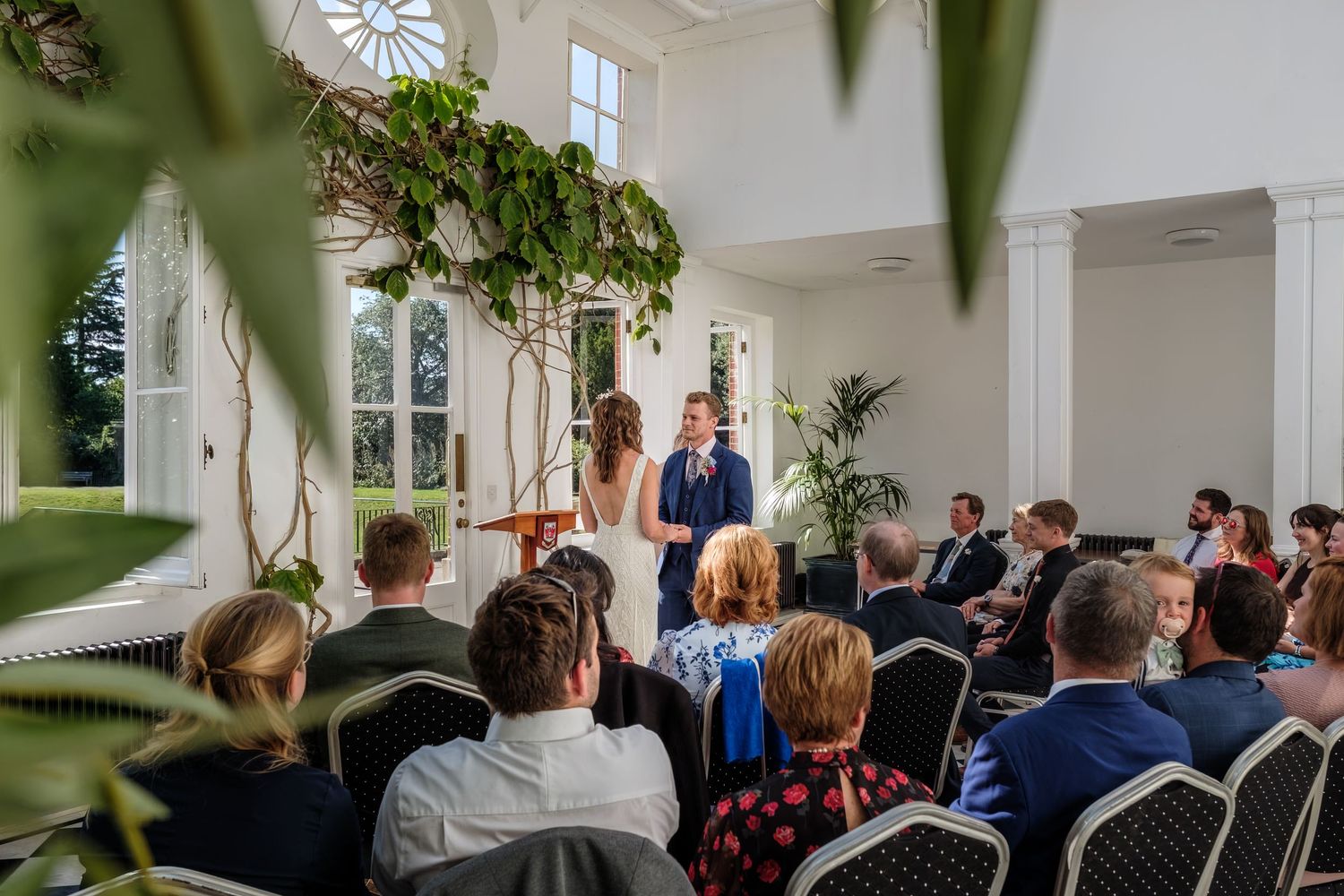 Elegant wedding ceremony in a bright white venue with guests seated as couple exchanges vows beneath hanging greenery.