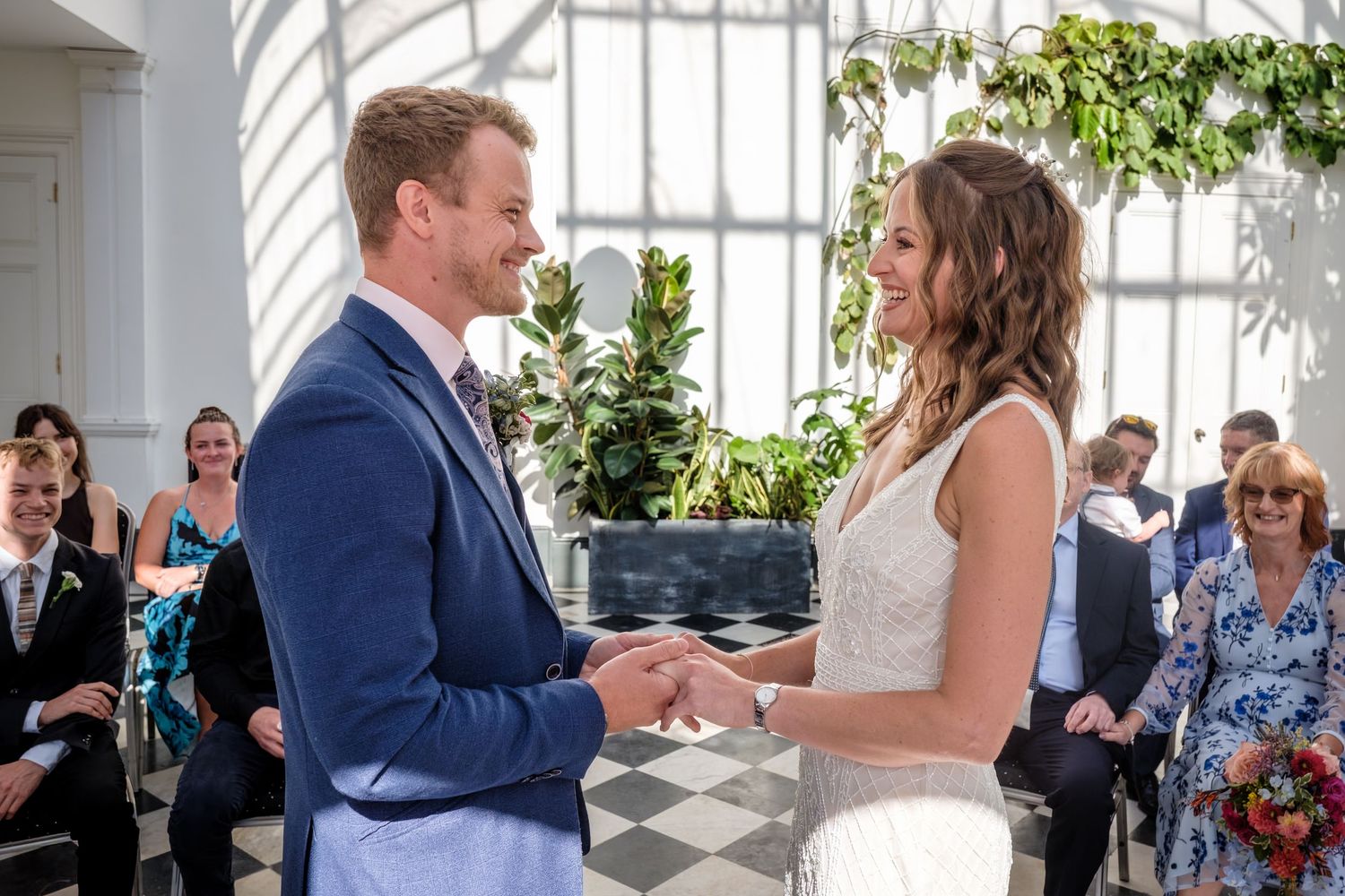 Couple exchanging vows at elegant wedding ceremony with guests seated and lush greenery backdrop.