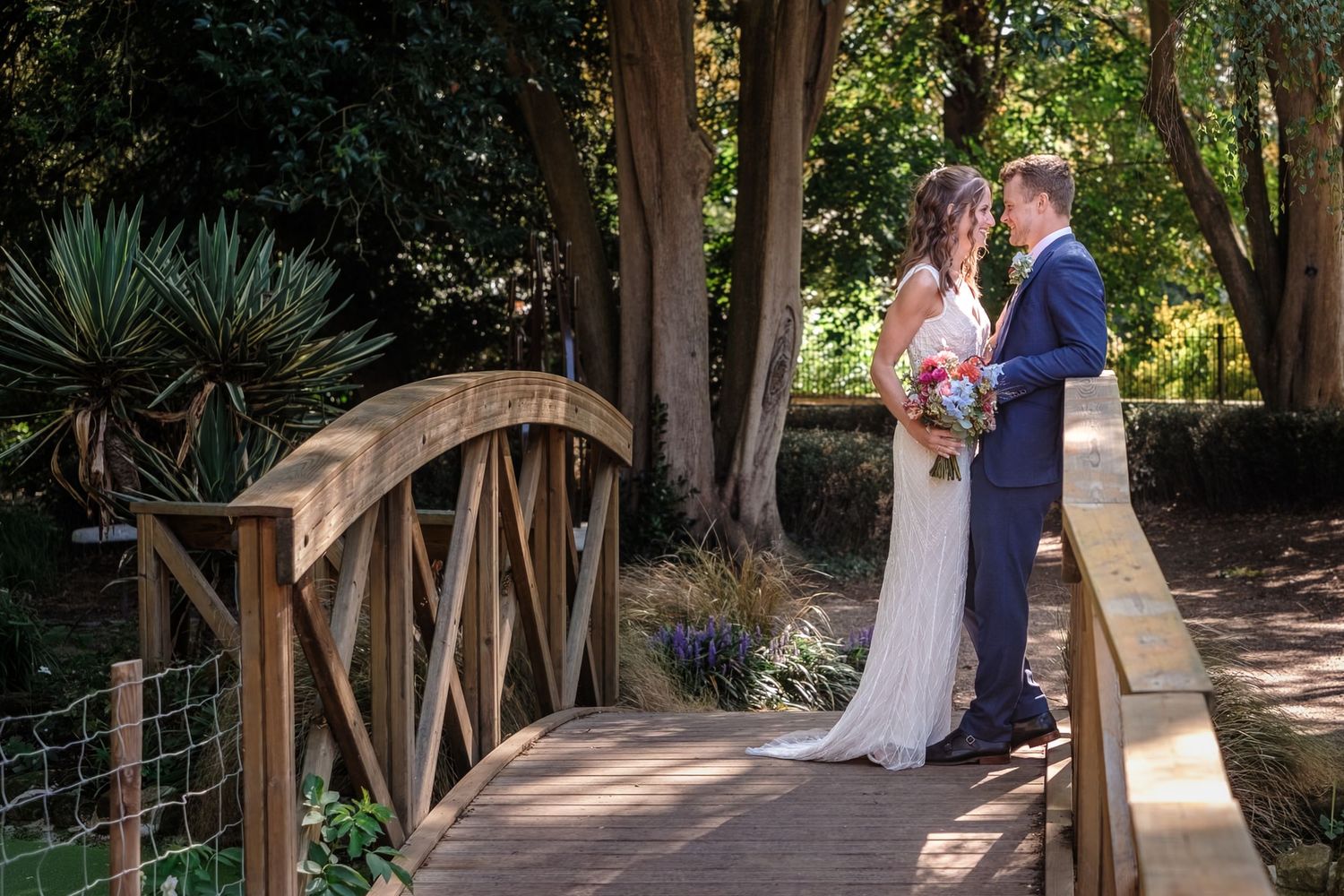 Bride and groom share a romantic moment on a wooden garden bridge surrounded by lush greenery on their wedding day.