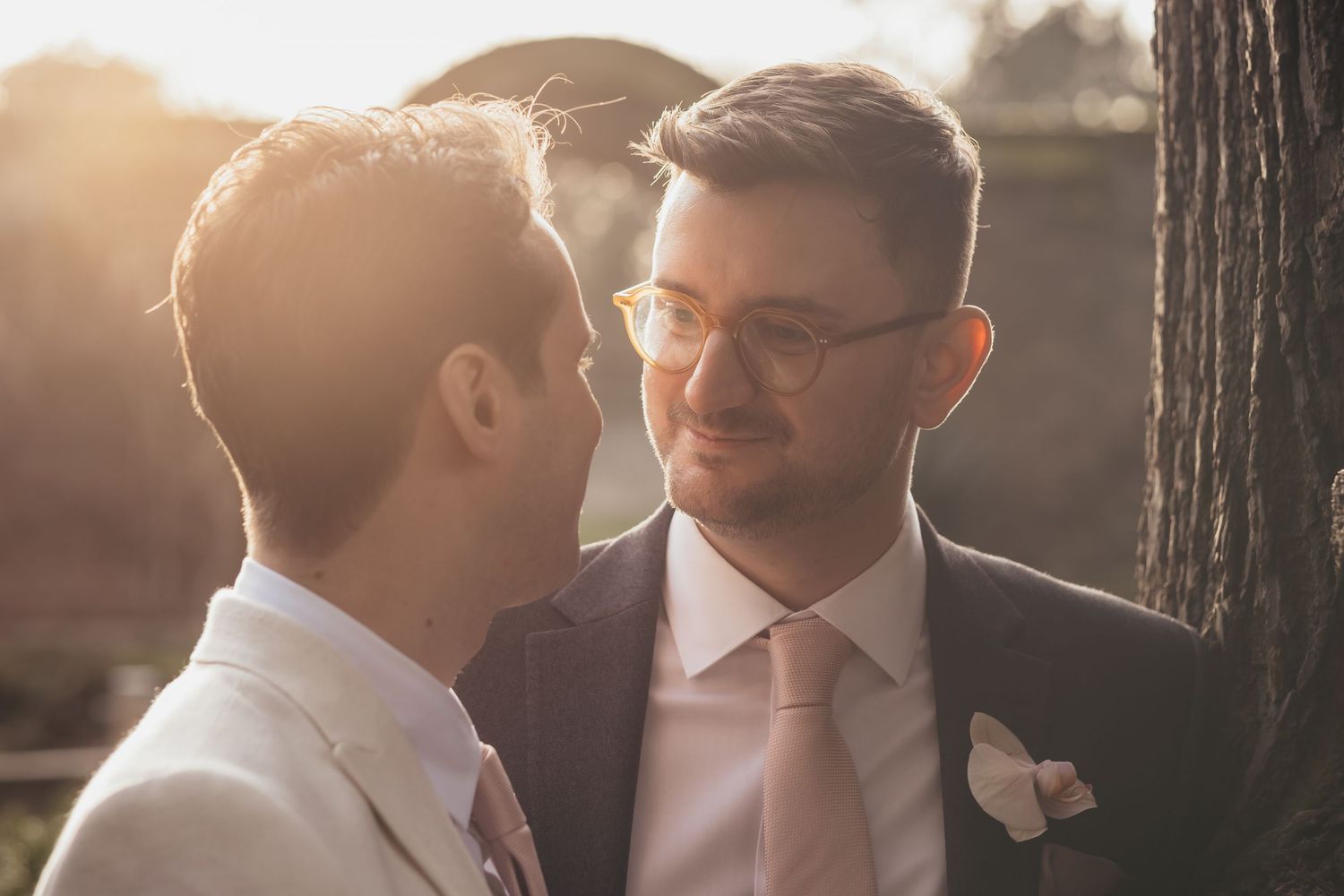 Two grooms share a romantic golden hour moment outdoors, one wearing glasses and a suit with a pink tie and boutonniere.