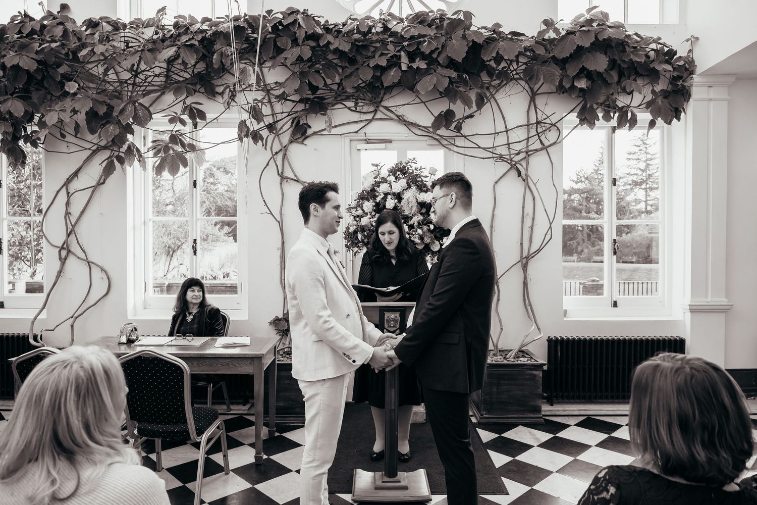 Two grooms hold hands at wedding altar adorned with floral arch and vines in elegant black and white checkered venue.